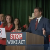 a man in a blue suit standing behind a podium with a sign that says "stop woke act"